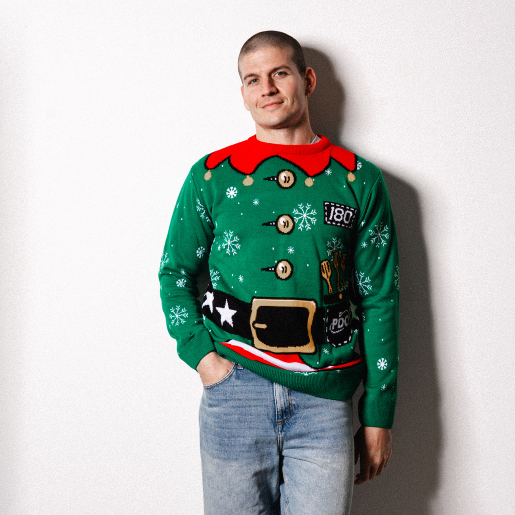 Wearing the Professional Darts Corporation PDC Elf Jumper, a person smiles slightly against a white wall, resting one hand in their jeans pocket.