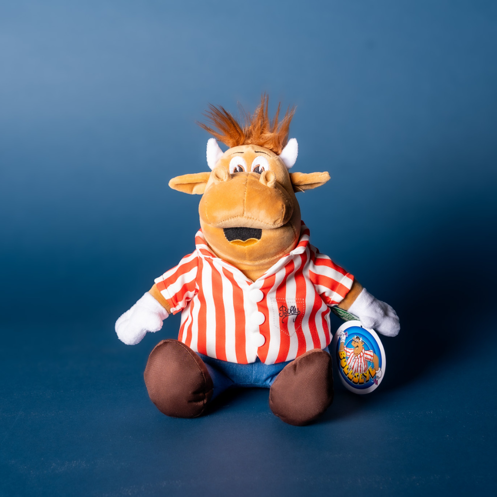 The Professional Darts Corporation Bullseye Plush, featuring brown fur, white horns, and a red-and-white striped shirt, sits against a blue background holding a tag with art matching the 9-inch cake pan design.