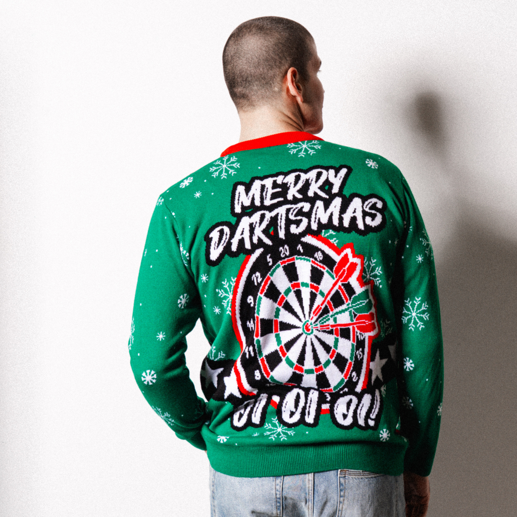 A man faces away, wearing the Professional Darts Corporation’s PDC Elf Jumper—a green sweater with white snowflakes, a large dartboard and red darts on the back, and “Merry Dartsmas” written across it.