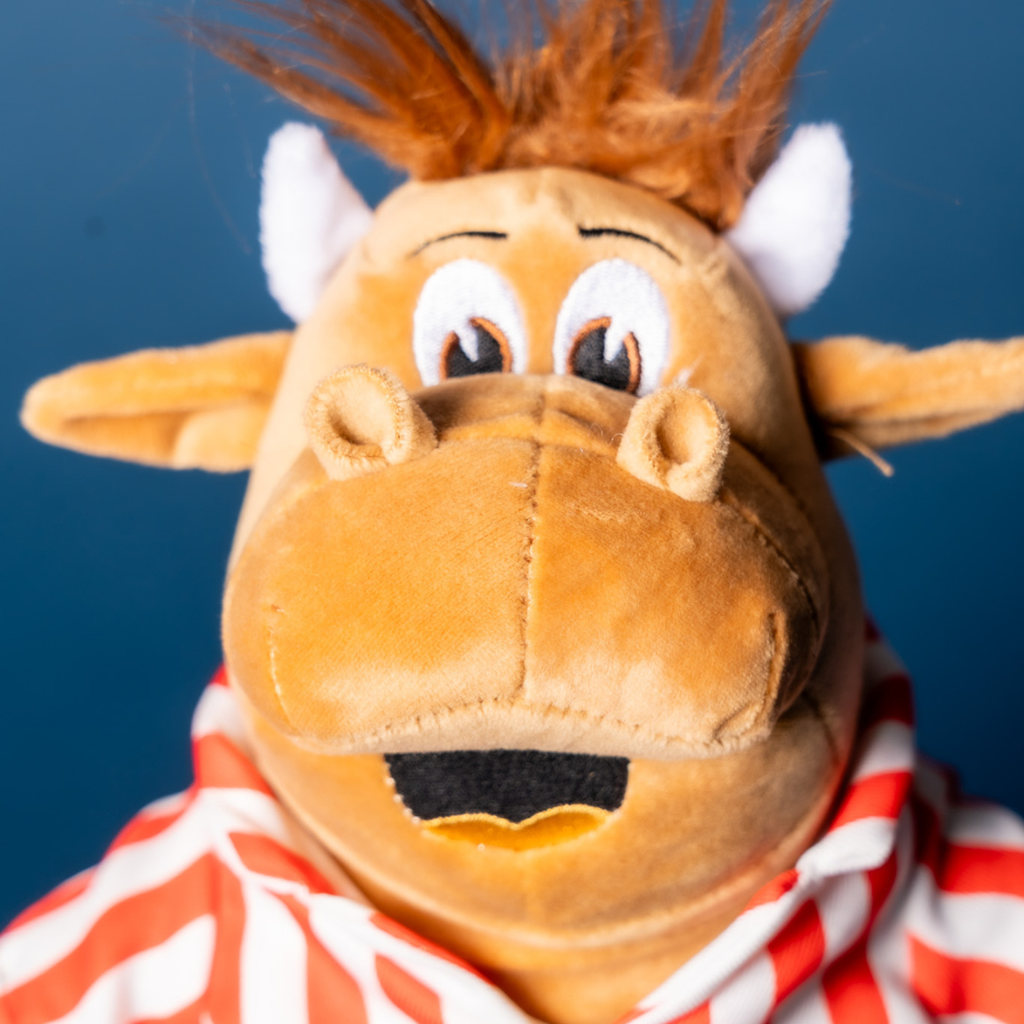 A close-up of the Bullseye Plush by Professional Darts Corporation—a cow with brown fur, white horns, big eyes, and a surprised look in a red and white striped outfit. A 9 inch cake pan on the blue background adds a playful kitchen vibe.